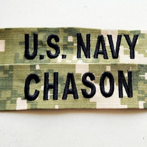 May include: Two embroidered fabric name tapes. The top tape reads "U.S. NAVY" in black block letters. The bottom tape reads "CHASON" in black block letters. The fabric has a digital camouflage pattern in shades of green, tan, and beige.