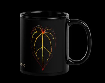 May include: Black ceramic mug with a colorful leaf design. The leaf is outlined in yellow and filled with red and orange hues. The mug has the word "STUDIO" printed on the bottom.