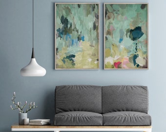 Puede incluir: Dos pinturas abstractas en tonos verdes, azules y amarillos apagados, colgadas en una pared azul claro sobre un sofá gris con patas de madera.