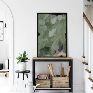 May include: An abstract painting in a black frame, featuring shades of green, white, and touches of purple and beige. The artwork is displayed on a wooden console table with a black metal frame, alongside wooden crates and a leather bag. A potted plant and a decorative metal pitcher are also visible.