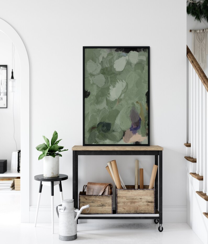 May include: An abstract painting in a black frame, featuring shades of green, white, and touches of purple and beige. The artwork is displayed on a wooden console table with a black metal frame, alongside wooden crates and a leather bag. A potted plant and a decorative metal pitcher are also visible.