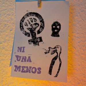 Op de afbeelding: Een witte papieren afdruk met zwarte en paarse inkt. De afdruk toont een vrouwelijk symbool met een vuist, een bivakmuts en een gestileerde figuur. De woorden "NI UNA MENOS" zijn in paars gedrukt. De afdruk wordt vastgehouden door een wasknijper.