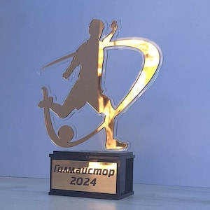 May include: A gold-colored trophy with a silhouette of a soccer player kicking a ball. The base of the trophy is black with a gold plate that reads "Голмайстор 2024".