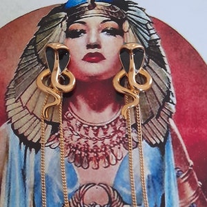 May include: A pair of gold-toned drop earrings featuring a stylised cobra head with black enamel detailing. Each earring has a long, delicate gold chain hanging from the base of the snake, creating a slender, dangling silhouette that catches the light.