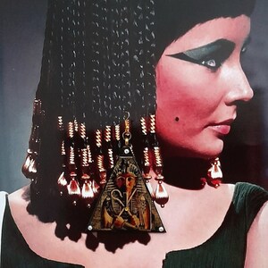 Może przedstawiać: Portret kobiety z ciemnymi włosami i dramatycznym makijażem oczu, ubranej w ciemnozieloną sukienkę i złotą biżuterię. Kolczyki są trójkątne z egipskim wzorem faraona. Widoczna złota bransoletka w kształcie węża.