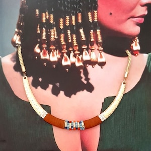 Puede incluir: Un collar dorado con un centro marrón y detalles coloridos, combinado con pendientes a juego. El collar se presenta sobre un fondo de una mujer con un vestido verde, con elaboradas trenzas y maquillaje.