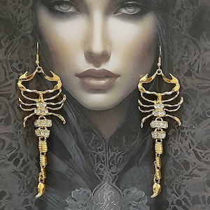 May include: Gold scorpion earrings with clear crystal detailing. The earrings are set against a portrait of a woman with dark hair and a detailed headpiece. The earrings have a hook closure.