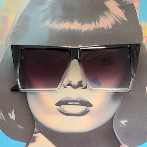 May include: A pair of black oversized sunglasses with a square frame and gradient lenses. The sunglasses are worn by a woman with short black hair.