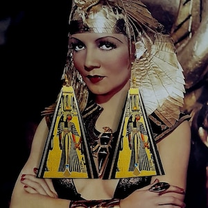 May include: Gold-toned Egyptian-themed earrings with a black border and a yellow background. The earrings feature a hieroglyphic design with a figure in a long gown. The woman in the image is wearing a gold headpiece and jewelry.