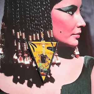 Może przedstawiać: Trójkątny kolczyk z motywem egipskim. Kolczyk ma żółte tło z hieroglifami i postaciami, obramowane czarną obwódką. Wisi na złotym haczyku. Obraz jest ustawiony na tle kobiety.