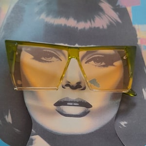 May include: A woman wearing large, angular, yellow-tinted sunglasses with a green frame. The sunglasses have a sharp, geometric shape.