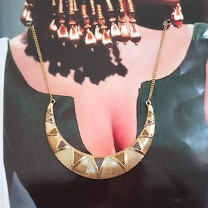 Könnte beinhalten: Eine goldfarbene Halskette mit einem halbmondförmigen Anhänger mit geometrischen Mustern. Das Bild zeigt auch eine Frau mit dunklem Haar, aufwendigem Kopfschmuck und dramatischem Augen-Make-up, das an das alte Ägypten erinnert.