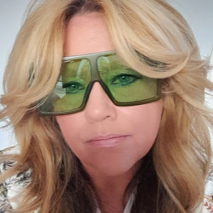 May include: A woman with long blonde hair wearing green tinted sunglasses. She is wearing a white shirt with a floral pattern.