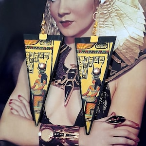 Puede incluir: Pendientes dorados de temática egipcia con forma triangular, con jeroglíficos y una ilustración de una figura egipcia. Los pendientes se llevan con joyas doradas a juego, incluyendo un brazalete y un anillo. La imagen tiene una estética vintage.