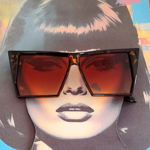 May include: A pair of brown and black oversized sunglasses with a geometric frame. The sunglasses have a gradient lens that is darker at the top and lighter at the bottom.