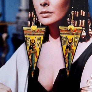 May include: A portrait of a woman wearing large, triangular earrings with an Egyptian theme. The earrings are gold, yellow, and black, featuring hieroglyphs and a depiction of an Egyptian figure. The woman has dark hair and dramatic blue eye makeup.