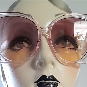 May include: Clear-framed sunglasses with gradient lenses. The lenses transition from a dark pink at the top to a yellow-orange at the bottom. The sunglasses are displayed on a mannequin head with painted features.