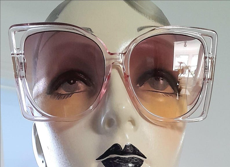 May include: Clear-framed sunglasses with gradient lenses. The lenses transition from a dark pink at the top to a yellow-orange at the bottom. The sunglasses are displayed on a mannequin head with painted features.
