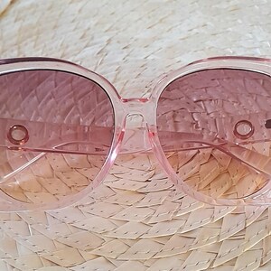May include: Pink square-framed sunglasses with gradient lenses. The frames are a transparent pink, and the lenses transition from a darker pink at the top to a lighter peach color at the bottom. The sunglasses are resting on a woven straw surface.