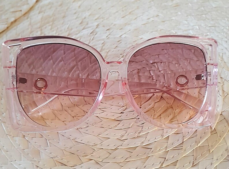 May include: Pink square-framed sunglasses with gradient lenses. The frames are a transparent pink, and the lenses transition from a darker pink at the top to a lighter peach color at the bottom. The sunglasses are resting on a woven straw surface.