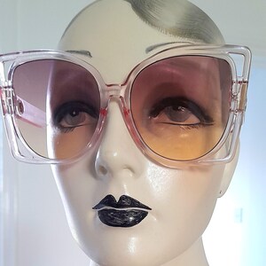 May include: Clear-framed sunglasses with gradient lenses. The lenses transition from a dark pink at the top to a yellow-orange at the bottom. The sunglasses have a square shape with a slight cat-eye design.
