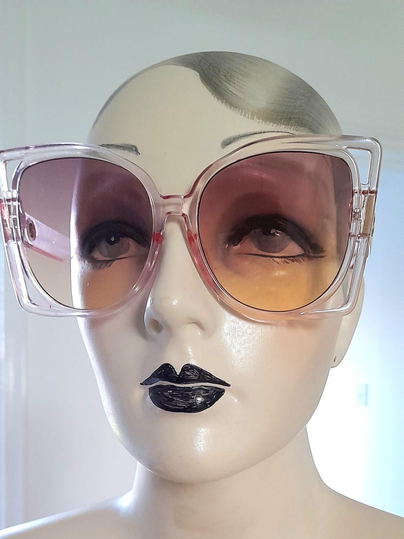 May include: Clear-framed sunglasses with gradient lenses. The lenses transition from a dark pink at the top to a yellow-orange at the bottom. The sunglasses have a square shape with a slight cat-eye design.