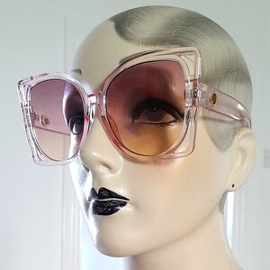 May include: Oversized, clear pink sunglasses with gradient lenses transitioning from purple to orange. The square frames have a unique, angular design. Gold-colored accents are visible on the temples. The sunglasses are displayed on a mannequin.