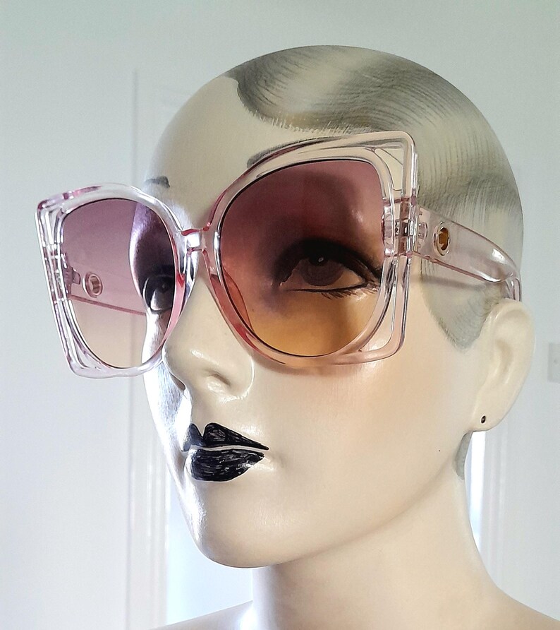 May include: Oversized, clear pink sunglasses with gradient lenses transitioning from purple to orange. The square frames have a unique, angular design. Gold-colored accents are visible on the temples. The sunglasses are displayed on a mannequin.