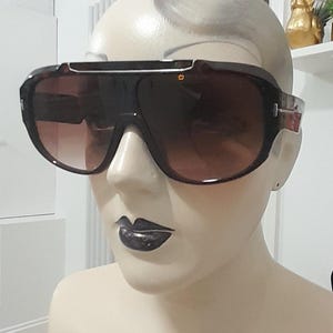 Puede incluir: Un par de gafas de sol grandes, marrones y envolventes con una montura negra. Las gafas de sol tienen una lente degradada, con un tono más oscuro en la parte superior y un tono más claro en la parte inferior.