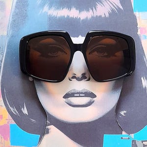 May include: A black and white illustration of a woman wearing large, black, square-shaped sunglasses with brown lenses.