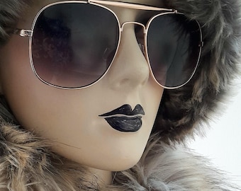 May include: Rose gold aviator sunglasses with dark gradient lenses. The sunglasses are set against a backdrop of a mannequin wearing a faux fur hat and collar. The mannequin has dark lipstick.