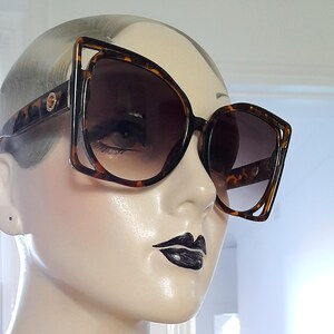 May include: Large, square-framed sunglasses with a tortoiseshell pattern. The sunglasses have a unique open-frame design on the outer edges and dark gradient lenses. The frames are brown and black, and the lenses are a dark brown color.
