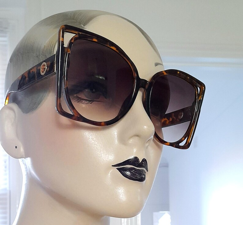 May include: Large, square-framed sunglasses with a tortoiseshell pattern. The sunglasses have a unique open-frame design on the outer edges and dark gradient lenses. The frames are brown and black, and the lenses are a dark brown color.