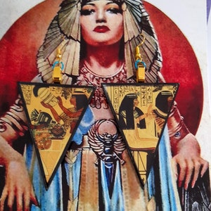 Puede incluir: Pendientes triangulares con temática egipcia. Los pendientes presentan ganchos de metal dorado y bordes negros. El diseño principal muestra jeroglíficos e ilustraciones de figuras egipcias en tonos dorados, marrones y negros. Los pendientes están sobre una portada de revista vintage.