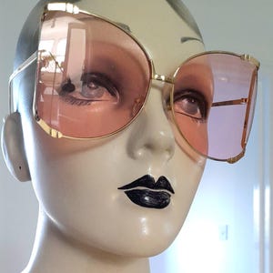 Könnte beinhalten: Sonnenbrille mit goldfarbenem Rahmen und großen, abgerundeten rosa Gläsern. Die Sonnenbrille hat ein Retro-Design in Übergröße. Das Bild zeigt die Sonnenbrille auf einem Schaufensterpuppenkopf mit schwarzem Lippenstift.