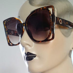 May include: Oversized sunglasses with a tortoiseshell frame and gradient lenses. The sunglasses have a square shape with a unique cut-out design on the outer corners. The lenses transition from a dark brown at the top to a lighter shade at the bottom.