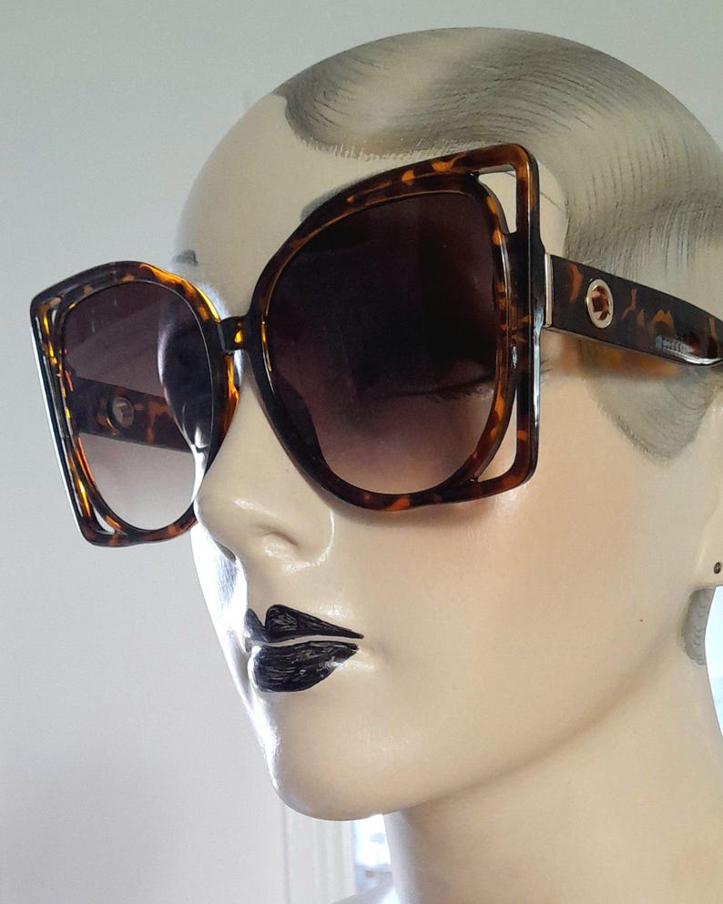 May include: Oversized sunglasses with a tortoiseshell frame and gradient lenses. The sunglasses have a square shape with a unique cut-out design on the outer corners. The lenses transition from a dark brown at the top to a lighter shade at the bottom.