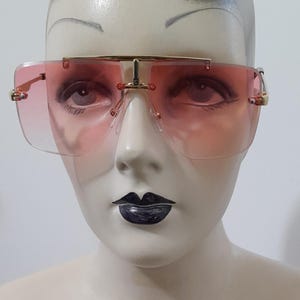May include: Pink-tinted sunglasses with a gold-coloured metal frame. The sunglasses have a square shape and a bridge that connects the two lenses. The lenses are a gradient pink colour, fading from a darker shade at the top to a lighter shade at the bottom.