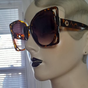 May include: Oversized sunglasses with a tortoiseshell pattern frame and dark gradient lenses. The sunglasses have a unique, angular design with a gold accent on the temples. The lenses transition from dark at the top to lighter at the bottom.