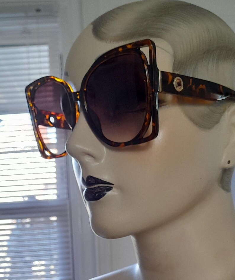 May include: Oversized sunglasses with a tortoiseshell pattern frame and dark gradient lenses. The sunglasses have a unique, angular design with a gold accent on the temples. The lenses transition from dark at the top to lighter at the bottom.