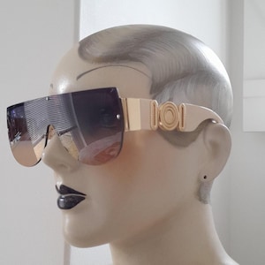 May include: Oversized shield sunglasses featuring a single continuous dark gradient lens and a flat top edge. The temples are light beige with a prominent gold-tone circular emblem at the hinge, connecting to a wide, smooth arm design.