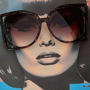 May include: Oversized sunglasses with a tortoiseshell frame and gradient lenses. The sunglasses are set against a pop art style illustration of a woman's face, featuring dark hair and bold makeup. The frames have a distinctive, angular design.
