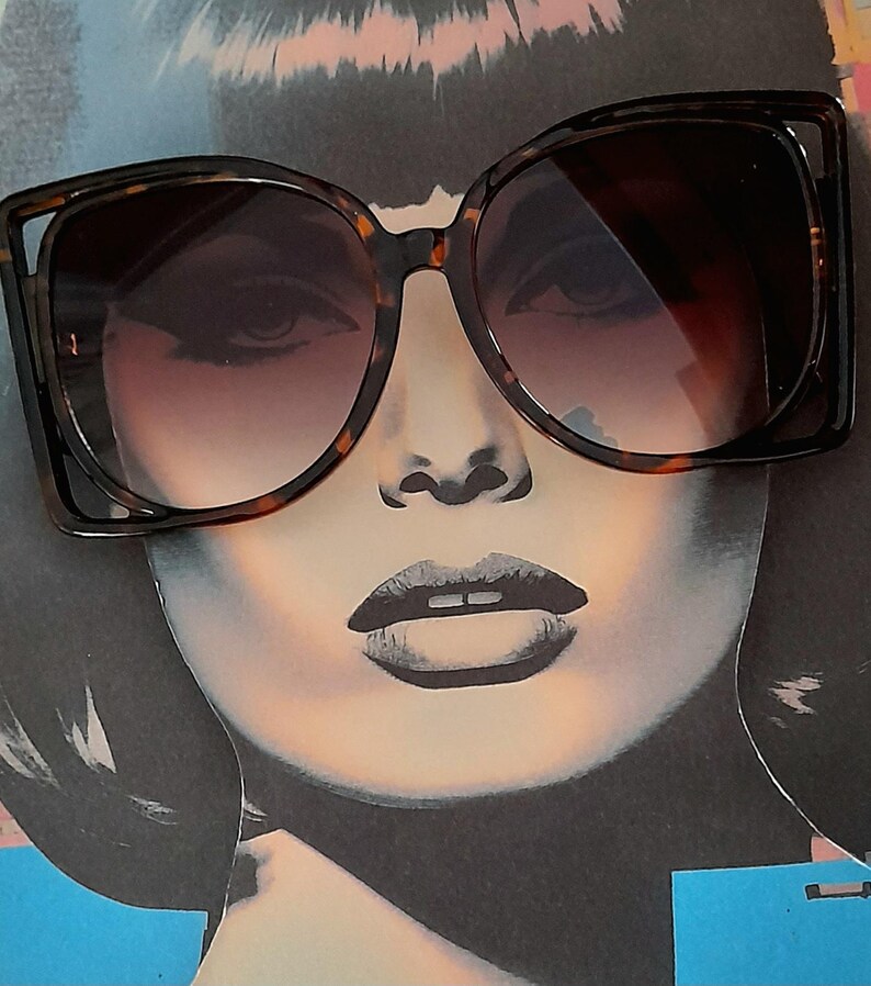 May include: Oversized sunglasses with a tortoiseshell frame and gradient lenses. The sunglasses are set against a pop art style illustration of a woman's face, featuring dark hair and bold makeup. The frames have a distinctive, angular design.