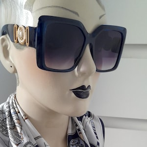 May include: Blue oversized square sunglasses with gold accents on the frame. The sunglasses have a gradient lens that fades from dark to light.