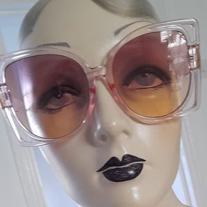 May include: Oversized square sunglasses with a clear frame and gradient lenses that transition from pink to orange. The sunglasses are displayed on a mannequin head with dark lipstick.