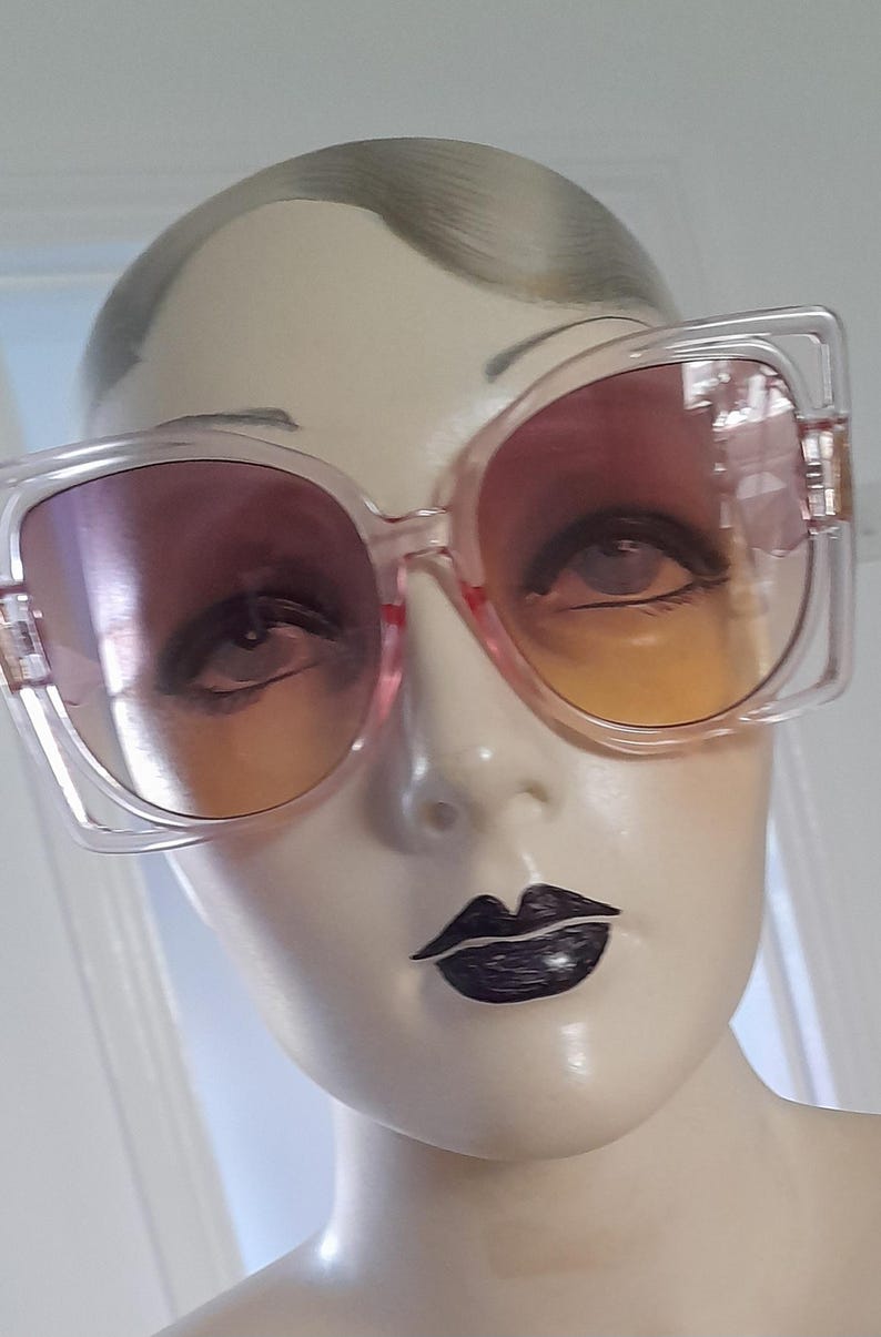 May include: Oversized square sunglasses with a clear frame and gradient lenses that transition from pink to orange. The sunglasses are displayed on a mannequin head with dark lipstick.