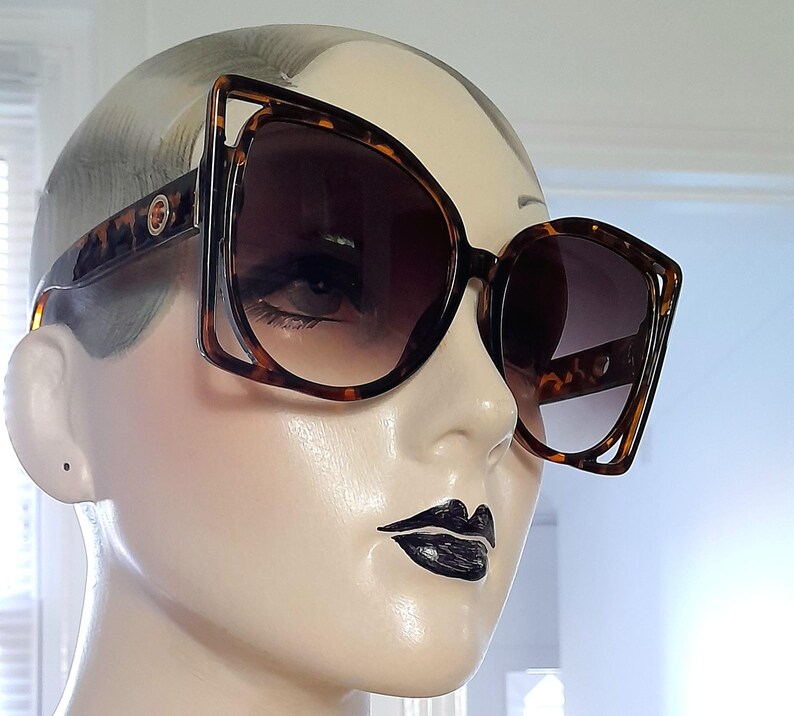 May include: Oversized sunglasses with a tortoiseshell pattern. The frames have a unique geometric shape with dark lenses. The sunglasses are displayed on a mannequin head.