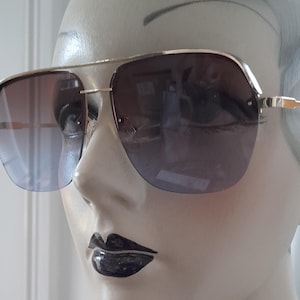 Puede incluir: Gafas de sol con montura dorada y lentes degradadas. Las gafas de sol tienen un diseño clásico de aviador con doble puente y un degradado sutil, de azul claro a marrón oscuro. Las gafas se exhiben en un maniquí.