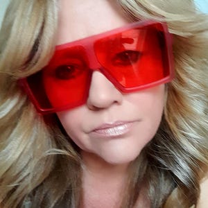 Può includere: Primo piano di una persona con grandi occhiali da sole rosso vivo. Gli occhiali da sole hanno una forma rettangolare e coprono completamente gli occhi. La persona ha i capelli biondi e la pelle chiara.