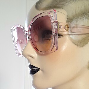 May include: Oversized pink sunglasses with a square frame and gradient lenses. The sunglasses have a light pink frame and temples with a gold-colored accent near the hinges. The lenses transition from a darker pink at the top to a lighter shade at the bottom.